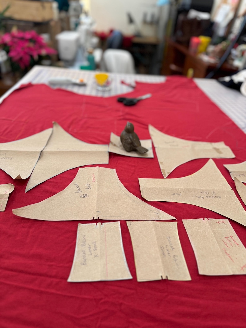 May include: A red fabric surface with multiple brown paper pattern pieces for sewing a garment. The pattern pieces are labeled with handwritten instructions, including "Bust Dart", "Back Neck", "Shoulder Piece", "Bust", "Back", and "Front".