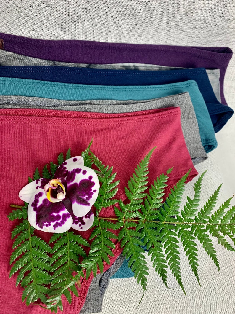 May include: A stack of six pairs of underwear in various colors, including purple, blue, teal, gray, and pink. A fern with a white orchid with purple accents is in the foreground.