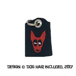 May include: Black fabric key chain with a silver key ring and a red embroidered dog head. The text "DESIGN &copy; DOG HAIR INCLUDED, 2017" is printed below the image.