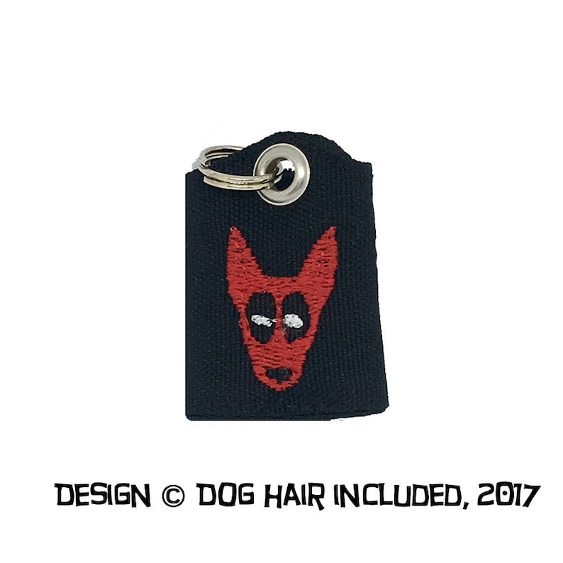 May include: Black fabric key chain with a silver key ring and a red embroidered dog head. The text "DESIGN &copy; DOG HAIR INCLUDED, 2017" is printed below the image.