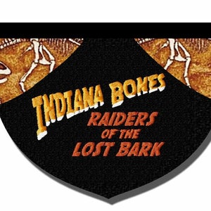 May include: A black patch with the text "Indiana Bones Raiders of the Lost Bark" in orange and yellow embroidery. The patch has a brown and orange background with a dinosaur skeleton design.