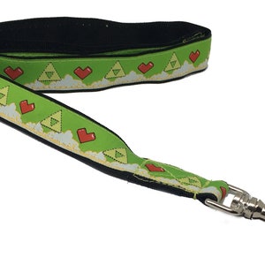 May include: Green dog lead with a repeating pattern of red hearts and a pixelated triangle design. The lead has a silver metal clip.