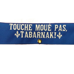 May include: Blue fabric patch with gold-colored metal fasteners. The patch has white embroidered text that reads "Touche moué pas, Tabarnak!" with a fleur-de-lis on either side of the text.