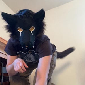 Puede incluir: Una máscara de animal similar a un lobo negro con pelaje texturizado, cubiertas de malla dorada para los ojos y marcas blancas debajo de los ojos. La máscara presenta orejas puntiagudas y un hocico con un pequeño detalle de nariz dorada.