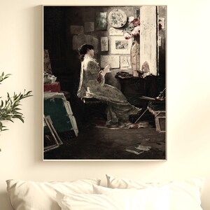 Könnte beinhalten: Gerahmtes Vintage-Gemälde, das eine Frau in einem Raum zeigt, möglicherweise ein Künstleratelier. Die Frau trägt ein helles Kleid. Der Raum verfügt über Kunstwerke, ein Fenster und einen Tisch mit Kunstutensilien. Die Farbpalette ist gedämpft mit dunklen Schatten.