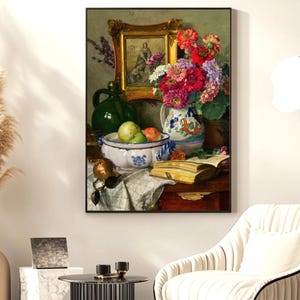 May include: A framed still life painting depicting a vase of vibrant flowers, a bowl of fruit, and an open book. A green jug and a decorative framed picture are also present. The painting is set on a table covered with a white cloth.