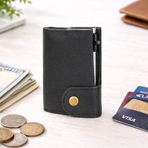 May include: A black leather wallet with a gold button closure. A silver pen is visible in the wallet's side pocket. The wallet is next to a stack of US dollar bills, coins, and credit cards.
