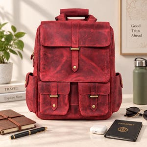 Pode incluir: Uma mochila de couro vermelho escuro com uma aba superior com fecho de fivela, dois bolsos frontais com botões de pressão e bolsos laterais. O material tem um acabamento texturizado e desgastado com padrões manchados em vermelho escuro.