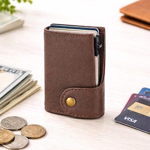 May include: A brown textured leather card holder wallet featuring a vertical orientation, a single brass-colored snap button closure, and an integrated mechanical card ejection mechanism visible at the top with several cards inserted inside.