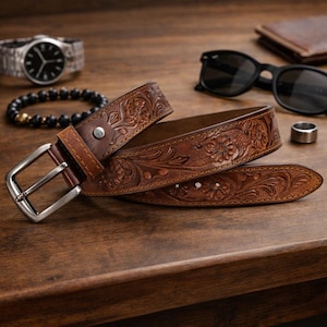 May include: A brown leather belt featuring an intricate floral embossed pattern and a silver-toned metal buckle. The belt is coiled and displays detailed craftsmanship with visible stitching along the edges and a single metal rivet near the buckle.