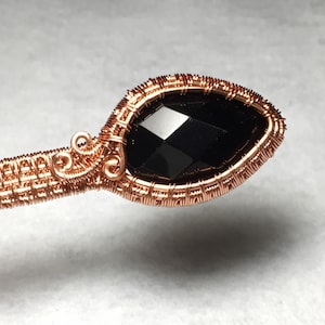 Puede incluir: Un colgante de piedra negra envuelto en alambre de cobre. El alambre est&aacute; tejido en un patr&oacute;n delicado alrededor de la piedra preciosa.