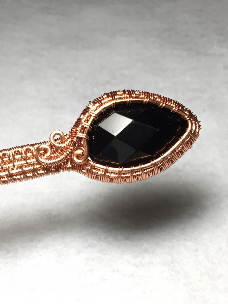 Puede incluir: Un colgante de piedra negra envuelto en alambre de cobre. El alambre est&aacute; tejido en un patr&oacute;n delicado alrededor de la piedra preciosa.