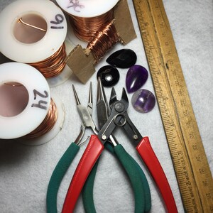 Puede incluir: Dos carretes de alambre de cobre, uno etiquetado como "2H" y el otro como "1H", con una regla de madera, dos alicates y varias piedras preciosas de amatista y negras.