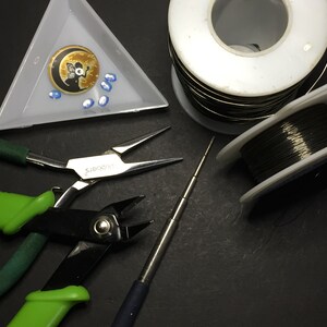 May include: A set of jewellery making tools including two pairs of pliers, a wire wrapping tool, two spools of wire, and a small tray with a moon charm and blue beads.