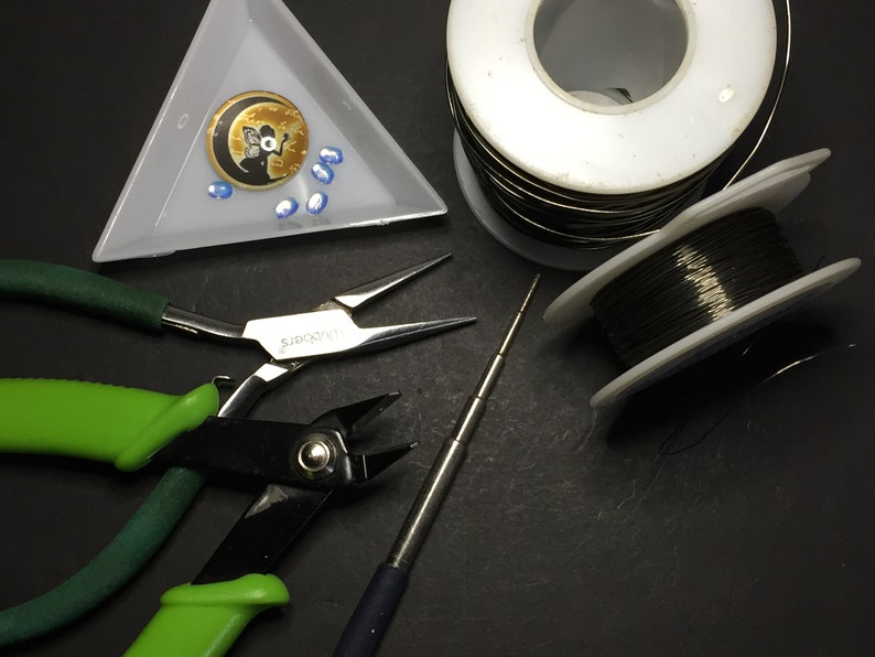 May include: A set of jewellery making tools including two pairs of pliers, a wire wrapping tool, two spools of wire, and a small tray with a moon charm and blue beads.