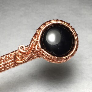 Puede incluir: Un colgante envuelto en alambre de cobre con una piedra preciosa negra en el centro. El colgante est&aacute; hecho con un dise&ntilde;o de alambre tejido.