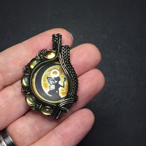 May include: A wire-wrapped pendant featuring a round, black and white image of a fairy with wings, sitting on a crescent moon. The image is surrounded by a wire frame with yellow iridescent stones.
