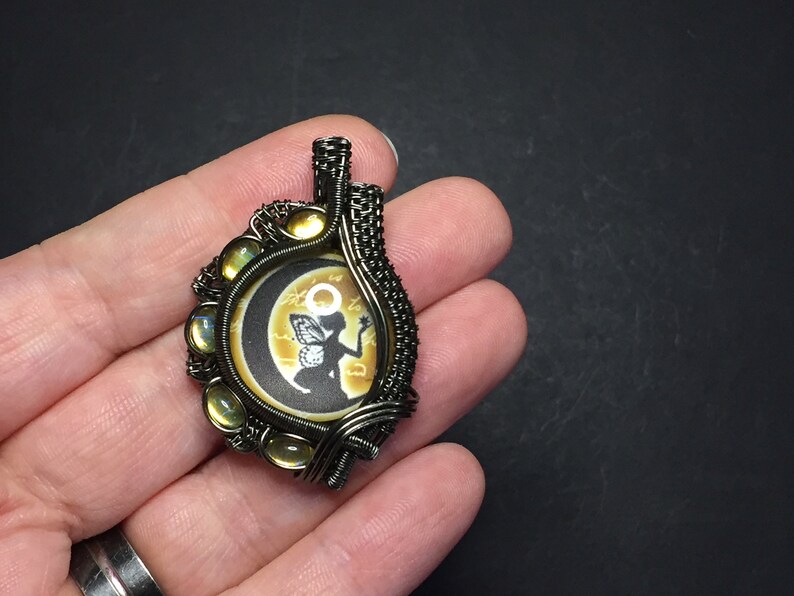 May include: A wire-wrapped pendant featuring a round, black and white image of a fairy with wings, sitting on a crescent moon. The image is surrounded by a wire frame with yellow iridescent stones.
