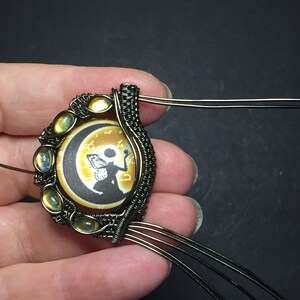 May include: A wire-wrapped pendant featuring a round cameo with a silhouette of a fairy holding a star, set against a crescent moon and a yellow background. The cameo is surrounded by a halo of iridescent yellow stones.