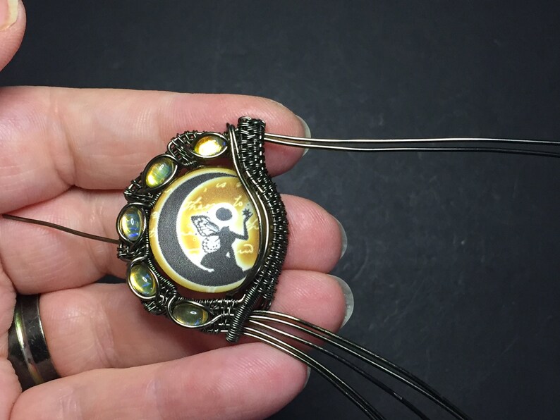 May include: A wire-wrapped pendant featuring a round cameo with a silhouette of a fairy holding a star, set against a crescent moon and a yellow background. The cameo is surrounded by a halo of iridescent yellow stones.