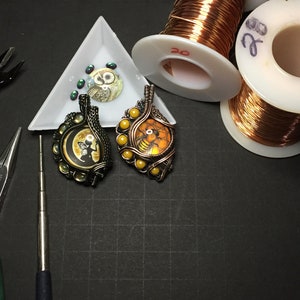 May include: Two wire wrapped pendants, one with a black and white owl and the other with a yellow and orange bee, are displayed on a black surface.  The pendants are surrounded by tools used for jewellery making, including wire, pliers, and a small tray of beads.