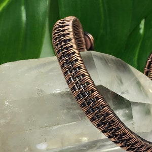 Puede incluir: Un brazalete tejido con alambre de cobre con un dise&ntilde;o trenzado. El brazalete se muestra sobre una superficie de cristal transparente.