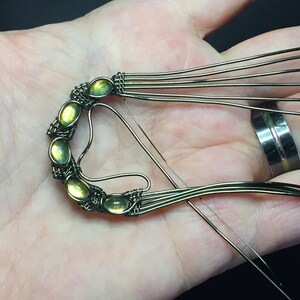 May include: A close-up of a wire-wrapped pendant featuring six green gemstones. The pendant is being held in a hand, and the wire is being wrapped around the stones.