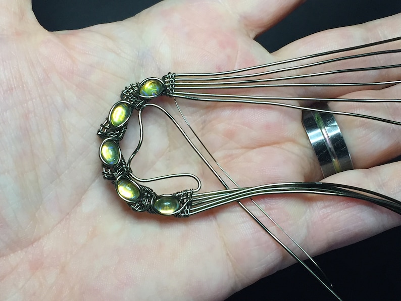 May include: A close-up of a wire-wrapped pendant featuring six green gemstones. The pendant is being held in a hand, and the wire is being wrapped around the stones.
