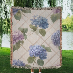 Puede incluir: Una manta tejida con un diseño floral de hortensias en tonos azules y morados. La manta tiene un fondo color crema con un patrón de cuadrícula y bordes con flecos. La manta se sostiene frente a una persona de pie sobre hierba verde.