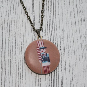 May include: A bronze-toned necklace with a round pendant featuring a cartoon soldier in a tall hat and uniform. The pendant is a light brown color. The necklace chain is also bronze-toned.