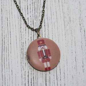 May include: A round, bronze-colored locket necklace with a detailed illustration of a toy soldier in a red uniform. The locket hangs from a delicate chain, creating a vintage aesthetic. The background is a light-colored, textured surface.