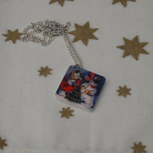May include: A silver chain necklace with a square pendant featuring a vintage illustration of two snowmen building a snowman in front of a blue and white winter scene.