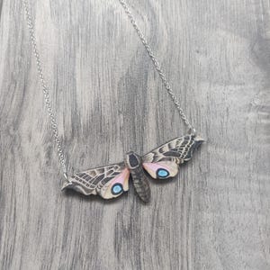 May include: A silver chain necklace with a wooden pendant shaped like a moth with pink and brown wings and blue eyes.