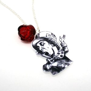 May include: A silver chain necklace with a red glass rose charm and a black and white illustration of the Mad Hatter from Alice in Wonderland. The Mad Hatter is wearing a top hat with the text "In this mad world" on it.