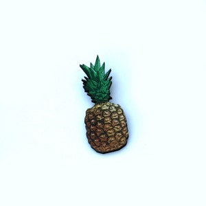 Könnte beinhalten: Eine hölzerne Ananas-Nadel mit grünem Oberteil und gelbem Körper. Die Ananas ist mit einem realistischen Design bemalt.