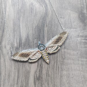 May include: A silver chain necklace with a large, detailed, wooden pendant shaped like a moth. The moth is painted with brown, white, and yellow markings.