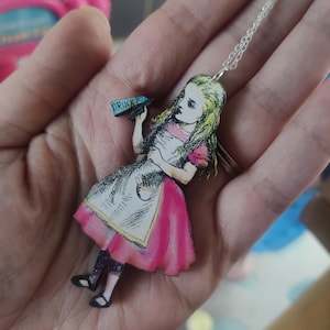 May include: Alice in Wonderland necklace featuring a wooden cutout of Alice holding a bottle that says "Drink Me". Alice is wearing a pink and white dress and has long blonde hair.