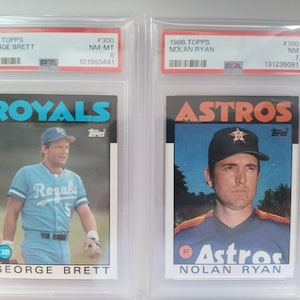 May include: Two 1986 Topps baseball cards, encased in protective holders. One features George Brett of the Royals, the other Nolan Ryan of the Astros. Both cards are in excellent condition, with clear images and vibrant colors.