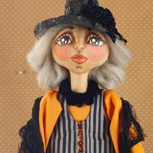 May include: A handmade witch doll with a pointed black hat and gray hair. The doll wears a black and white striped dress, an orange jacket, and a black shawl. The dress features a printed design with letters and illustrations.