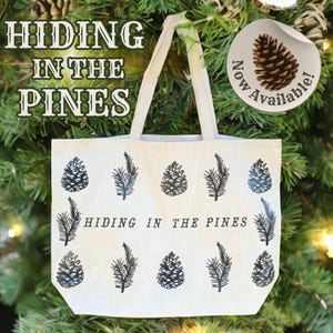 May include: A white canvas tote bag with black pine tree and pine cone illustrations. The bag has the text "Hiding in the Pines" printed on it. A sticker on the bag says "Now Available!" with a pine cone illustration.