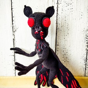 Könnte beinhalten: Eine schwarze gehäkelte Mothman-Figur mit roten Augen und Flügeln. Die Figur steht auf einer Holzoberfläche. "Mothman Crochet pattern pdf" steht in rosa Schrift am unteren Rand des Bildes.