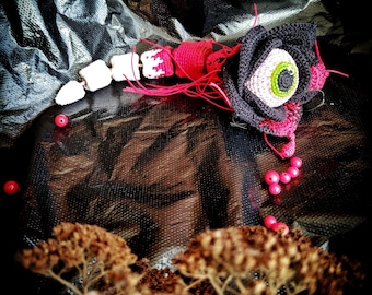 May include: Crochet pattern PDF for a black and pink amigurumi rose with a green eye. The rose is lying on a black surface with red beads scattered around it.