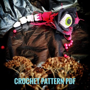 Puede incluir: Patrón de crochet PDF para una rosa amigurumi negra y rosa con un ojo verde. La rosa está sobre una superficie negra con cuentas rojas esparcidas a su alrededor.