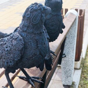 May include: Two black crow sculptures with detailed crochet-like texture are perched on a wooden park bench. The sculptures have realistic features, including beaks and feet, and are set against a blurred outdoor background.