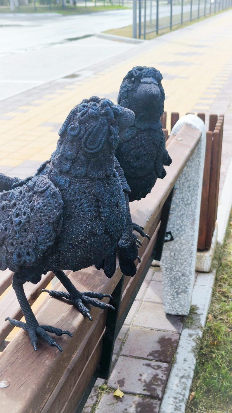 May include: Two black crow sculptures with detailed crochet-like texture are perched on a wooden park bench. The sculptures have realistic features, including beaks and feet, and are set against a blurred outdoor background.