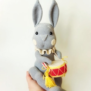 May include: A grey crocheted bunny doll with long ears, holding a small drum. The drum is red and yellow, with a white top. The doll has a white ruffled collar and blush-coloured cheeks. Text at the top reads "crochet pattern pdf in english."