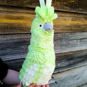 May include: A handmade, plush parrot in shades of green and white. The bird has a textured, feathery appearance, a gray beak, and black claws. The parrot is held in a person's hands against a wooden background. The parrot's height is approximately 12 inches.