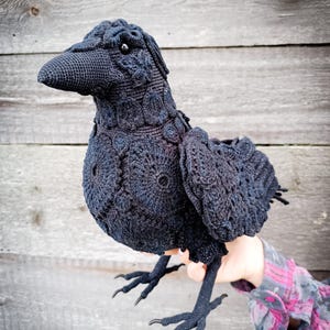 May include: A handcrafted, black crochet raven with detailed feather and eye features. The bird has a textured appearance and is held against a wooden background. The raven's legs and beak are also crocheted.
