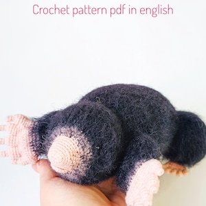 Puede incluir: Un peluche de topo hecho a mano con ganchillo en gris oscuro con patas y nariz de color rosa claro. El texto "Crochet pattern pdf in english" se muestra en rosa.