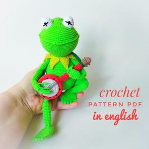Op de afbeelding: Groene gehaakte kikker amigurumi die een rode en witte banjo vasthoudt. Haakpatroon PDF in het Engels.